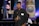 A person stands indoors holding a camera, looking directly at the viewer. They are wearing a black shirt and have a badge around their neck. Behind them, there are various banners with text and logos displayed on a stage, with blue and purple lighting adding color to the scene.