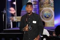 A person stands indoors holding a camera, looking directly at the viewer. They are wearing a black shirt and have a badge around their neck. Behind them, there are various banners with text and logos displayed on a stage, with blue and purple lighting adding color to the scene.
