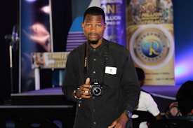 A person stands indoors holding a camera, looking directly at the viewer. They are wearing a black shirt and have a badge around their neck. Behind them, there are various banners with text and logos displayed on a stage, with blue and purple lighting adding color to the scene.