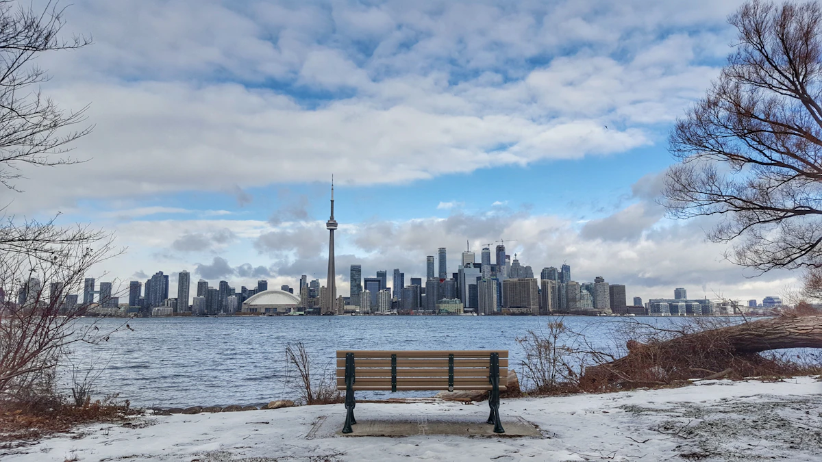 The Toronto skyline across the harbour on a snowy winter day, a park bench and snow-dusted shoreline in the foreground