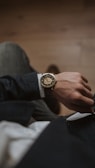 A close-up view of a person's wrist adorned with an elegant analog watch. The watch features a gold case and a visible gear mechanism, emphasizing its intricate design. The person is dressed in a dark-textured suit jacket and white shirt, with their hand resting on their lap.