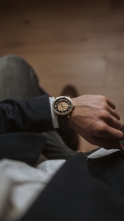 A close-up view of a person's wrist adorned with an elegant analog watch. The watch features a gold case and a visible gear mechanism, emphasizing its intricate design. The person is dressed in a dark-textured suit jacket and white shirt, with their hand resting on their lap.