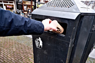 A hand is placing a disposable cup into a black outdoor trash bin on a cobblestone street. The trash bin is labeled with a graphic of a person throwing trash.