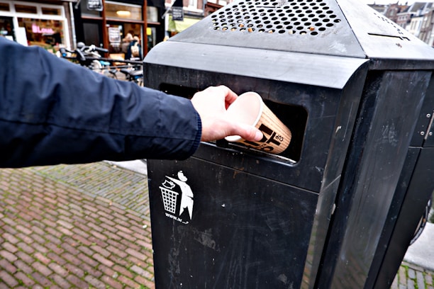 A hand is placing a disposable cup into a black outdoor trash bin on a cobblestone street. The trash bin is labeled with a graphic of a person throwing trash.