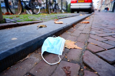 A disposable face mask lies on the cobblestone pavement next to a curb, surrounded by dried autumn leaves. A parked car and bicycles are visible in the blurry background on the street.