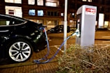 A black electric vehicle is connected to a charging station on a street, with a blue cable linking the car to the unit. The setting is an urban area at night, with surrounding buildings and illuminated windows. The paving is made of bricks, and there's a bush visible in the foreground.