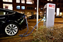 A black electric vehicle is connected to a charging station on a street, with a blue cable linking the car to the unit. The setting is an urban area at night, with surrounding buildings and illuminated windows. The paving is made of bricks, and there's a bush visible in the foreground.