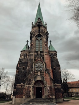 A large, ornate Gothic-style church with a tall central spire and two smaller ones on either side. The facade features intricate brickwork and stained glass windows, with a large circular window above the entrance. The surrounding area includes bare trees and a few buildings.