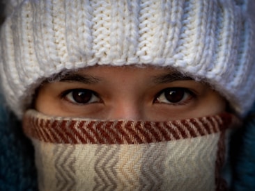 A person is wearing a warm, knitted white hat that covers part of the forehead and a thick, patterned scarf wrapped around the face, leaving only the eyes visible. The rich texture of the knitwear suggests a cozy and warm setting.