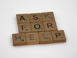 Wooden Scrabble tiles spell out the phrase 'ASK FOR HELP' arranged neatly on a white surface. Each tile shows a letter and a number indicating its point value in the game.