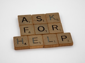 Wooden Scrabble tiles spell out the phrase 'ASK FOR HELP' arranged neatly on a white surface. Each tile shows a letter and a number indicating its point value in the game.