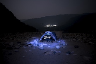 A tent is set up in a rocky and barren landscape under a dark sky. The tent is illuminated, and the word 'DAWKI' is visible on it. Distant lights from a village or town are seen in the background, nestled among shadowy hills.
