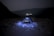 A tent is set up in a rocky and barren landscape under a dark sky. The tent is illuminated, and the word 'DAWKI' is visible on it. Distant lights from a village or town are seen in the background, nestled among shadowy hills.
