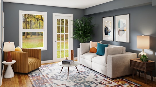 A modern living room with a cozy vibe features a large beige sofa adorned with colorful cushions, including teal and orange. There's a patterned area rug covering the wooden floor and a small wooden coffee table in the center. A stylish armchair is positioned next to a round side table with a lamp, near large windows that give a view of the outdoors. The walls are painted in a muted blue shade, and the room is elegantly decorated with minimalistic artwork and a large potted plant.