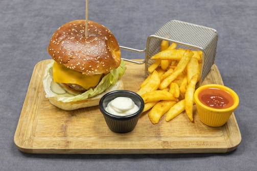A wooden board displays a sesame seed burger with cheese, lettuce, and a slice of onion. Alongside is a small metal basket with golden crispy fries, accompanied by two small pots containing mayonnaise and ketchup.