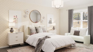 A cozy bedroom with a contemporary design featuring a large bed with textured bedding and several cushions. A round mirror is mounted on the wall above the headboard, flanked by decorative flowers. Two bedside tables with lamps add symmetry, and a plush white chair with pillows sits near the window. Natural light filters through gray curtains and white blinds, complementing the calming color palette of creams and grays.