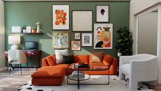 A well-decorated living room with a modern and colorful design. A green accent wall features an eclectic gallery of framed art. An orange sectional sofa with assorted cushions is complemented by a white, patterned armchair. A round coffee table with books and decorative items stands on a light rug with dark spots. A small desk with a lamp and chair is positioned against the wall, with shelves holding books and plants above it. A tall plant is situated in the corner.