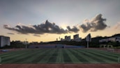 A sports field is situated in the foreground, with several people engaged in activities. The field is surrounded by a running track, and there are goalposts on either end. Behind the field, a line of modern buildings is visible, partially covering a hillside. Above, a dramatic cloud formation stretches across the sky, with the sun just below the horizon creating a silhouette effect.