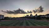 A sports field is situated in the foreground, with several people engaged in activities. The field is surrounded by a running track, and there are goalposts on either end. Behind the field, a line of modern buildings is visible, partially covering a hillside. Above, a dramatic cloud formation stretches across the sky, with the sun just below the horizon creating a silhouette effect.