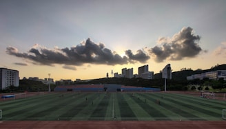 A sports field is situated in the foreground, with several people engaged in activities. The field is surrounded by a running track, and there are goalposts on either end. Behind the field, a line of modern buildings is visible, partially covering a hillside. Above, a dramatic cloud formation stretches across the sky, with the sun just below the horizon creating a silhouette effect.