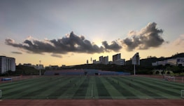 A sports field is situated in the foreground, with several people engaged in activities. The field is surrounded by a running track, and there are goalposts on either end. Behind the field, a line of modern buildings is visible, partially covering a hillside. Above, a dramatic cloud formation stretches across the sky, with the sun just below the horizon creating a silhouette effect.