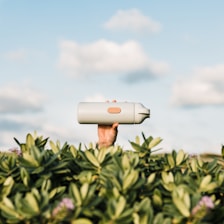 A hand is holding a light-gray water bottle above green foliage against a backdrop of a blue sky with fluffy clouds.