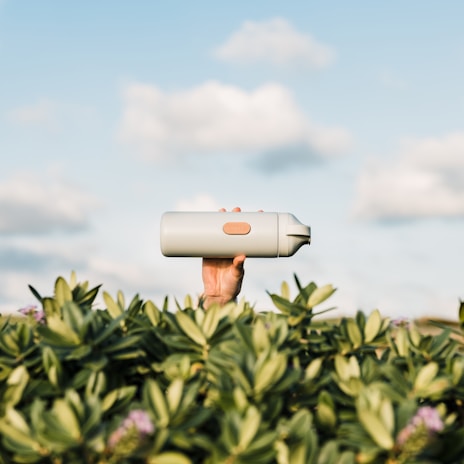 A hand is holding a light-gray water bottle above green foliage against a backdrop of a blue sky with fluffy clouds.