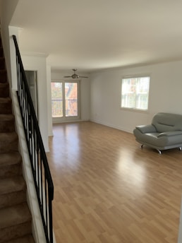 A spacious, unfurnished room with light wood flooring and white walls. There is a staircase with a dark railing on the left side leading upwards. Large windows on the far wall allow natural light to illuminate the space. A modern grey couch is positioned in the corner near the window. A ceiling fan is hanging from the ceiling above the open area.