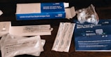 An assortment of COVID-19 self-test kits and materials, including test swabs, vials, instruction booklet, and packaging, are displayed on a wooden surface.