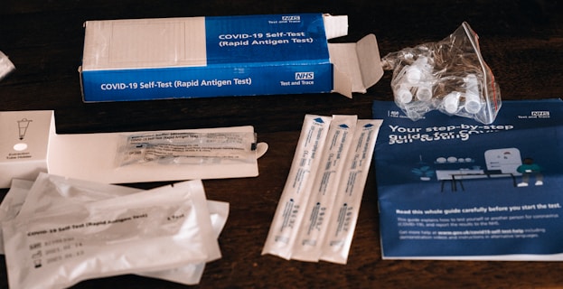 An assortment of COVID-19 self-test kits and materials, including test swabs, vials, instruction booklet, and packaging, are displayed on a wooden surface.