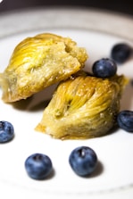 A piece of baklava is split open, showcasing its layers of golden, flaky pastry and filled with a pistachio blend. Surrounding the baklava are a few whole blueberries on a white plate, providing a contrast in color and texture.