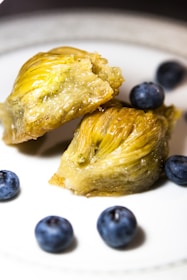 A piece of baklava is split open, showcasing its layers of golden, flaky pastry and filled with a pistachio blend. Surrounding the baklava are a few whole blueberries on a white plate, providing a contrast in color and texture.