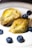 A piece of baklava is split open, showcasing its layers of golden, flaky pastry and filled with a pistachio blend. Surrounding the baklava are a few whole blueberries on a white plate, providing a contrast in color and texture.