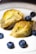 A piece of baklava is split open, showcasing its layers of golden, flaky pastry and filled with a pistachio blend. Surrounding the baklava are a few whole blueberries on a white plate, providing a contrast in color and texture.