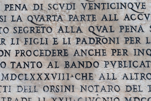 An aged stone surface with inscribed text in uppercase letters. The language appears to be Italian or Latin, with an ancient or classical style. The text is clearly visible and etched into the stone.
