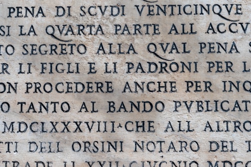 An aged stone surface with inscribed text in uppercase letters. The language appears to be Italian or Latin, with an ancient or classical style. The text is clearly visible and etched into the stone.