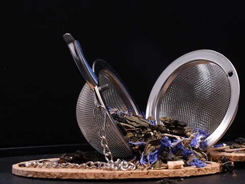 A silver tea infuser lies open on a circular cork mat, spilling out dried green tea leaves and small blue flowers. The infuser is made of fine mesh and has a chain attached. The background is a simple black, creating a contrast with the lighter colors of the objects.