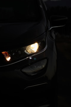 A close-up view of a car's front headlight illuminated in the dark. The light casts a soft glow, highlighting the sleek contours of the vehicle. The surrounding environment is dimly lit, adding to the contrast.
