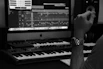 A music production setup is visible with a synthesizer keyboard in the foreground, a large computer monitor displaying audio editing software in the background, and a side view of a person wearing a watch, resting their arm near the keyboard.