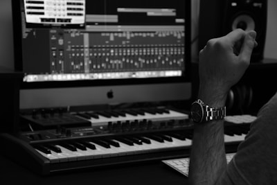 A music production setup is visible with a synthesizer keyboard in the foreground, a large computer monitor displaying audio editing software in the background, and a side view of a person wearing a watch, resting their arm near the keyboard.