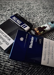The image contains multiple cigarette packs and a lighter. The cigarette packs include brands like Pall Mall and Winchester, with health warnings written on them in a foreign language. The lighter features a design with the silhouette of a cowboy and horse against a blue sky.