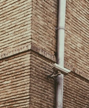 A corner of a brick building is featured with a surveillance camera mounted onto a vertical metal pipe. The bricks have a rough texture and are laid in a horizontal pattern. The camera is positioned at an angle, focused on the intersecting corner of the structure.