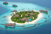 An aerial view of a tropical island resort featuring lush greenery, palm trees, and several thatched-roof villas surrounded by clear turquoise water. The island is bordered by white sandy beaches with a few isolated overwater bungalows in the surrounding sea.