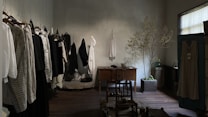 A dimly lit room features a row of clothing hung on hangers along the left wall. The garments, in shades of white, black, and beige, appear to be made of light, flowy fabrics. On the right side, a small potted tree adds a touch of greenery next to a wooden table holding several ceramic bowls. The floor is wooden, complementing the rustic yet minimalistic decor. A single window allows a soft, natural light to filter in, creating subtle shadows.