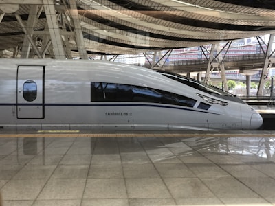 A sleek, modern high-speed train is parked at a station, reflecting sophisticated engineering with its streamlined design. The surrounding area features large metal beams and glass panels, typical of a train station environment.