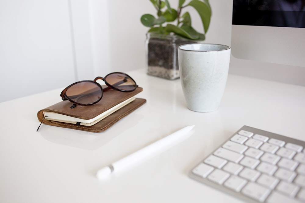 black framed sunglasses beside white apple keyboard and white ceramic mug