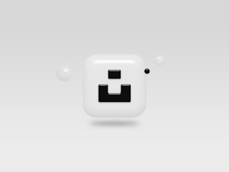 A glossy, rounded square icon with a bold black geometric design in the center, surrounded by small spherical objects on a minimalist gray background.