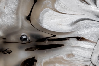 Swirling patterns of black and white create a marble-like texture, highlighted by a smooth, flowing design. Small bubbles and streaks of black are suspended within the cloudy, creamy surface, giving it an abstract and fluid appearance.