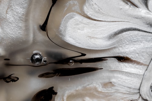 Swirling patterns of black and white create a marble-like texture, highlighted by a smooth, flowing design. Small bubbles and streaks of black are suspended within the cloudy, creamy surface, giving it an abstract and fluid appearance.