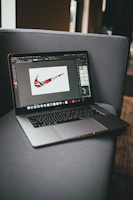 A laptop is placed on a gray couch, displaying a graphic design program with a colorful, patterned swoosh logo on the screen. The room is softly lit, with natural light coming from a window to the right.